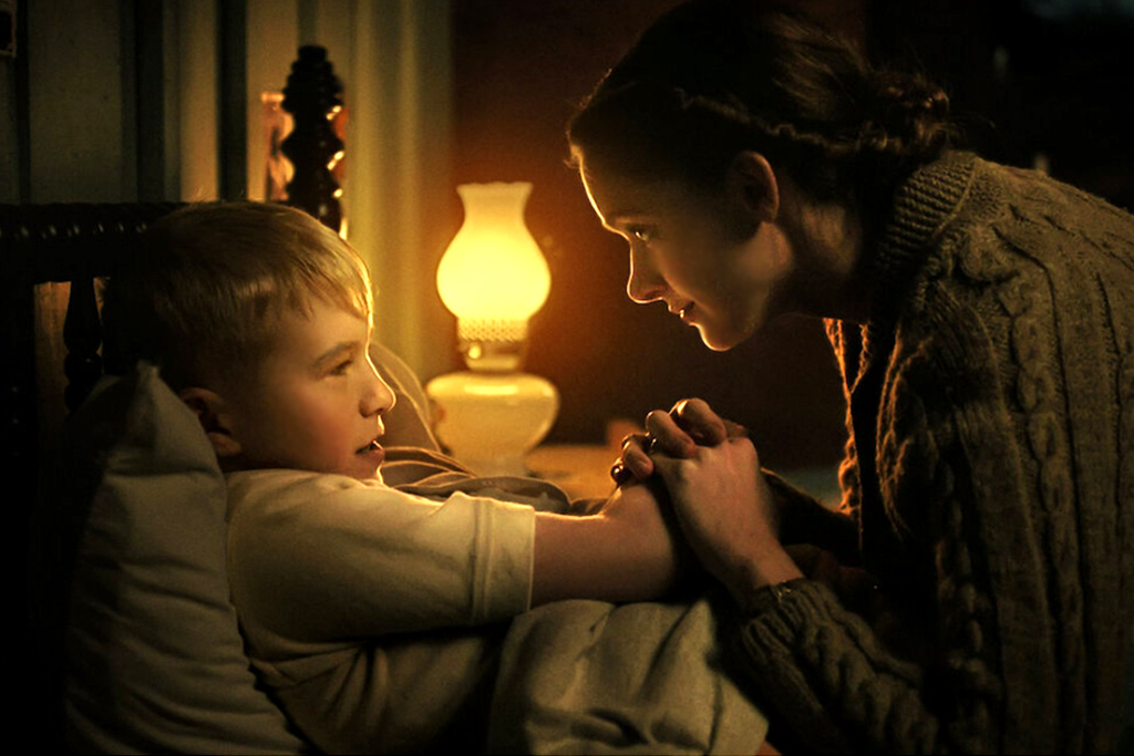 작은 조명만 켜져 있는 어두운 방. 한 여성이 침대에 비스듬히 누운 남자아이의 손을 두 손으로 감싸 잡고 눈을 마주치고 있다.