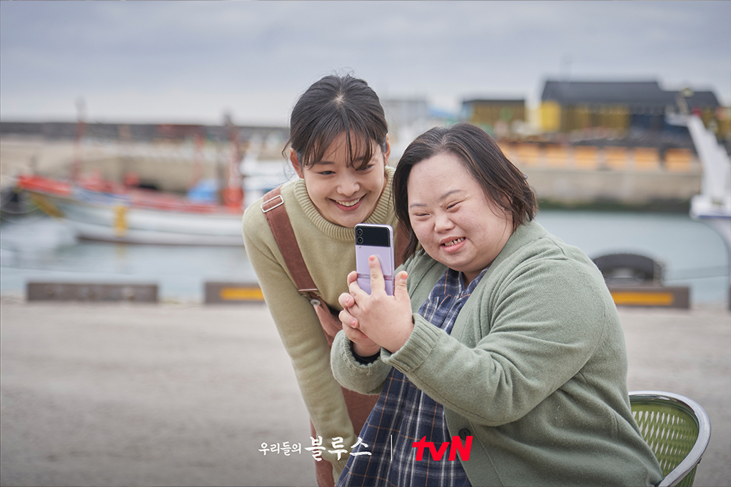 tvN 드라마 우리들의 블루스의 한장면. 청각장애 여성과 다운증후군 여성이 함께 휴대폰 화면을 보며 웃고 있다.