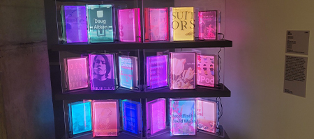 전시장. 여러 개 책 모양의 아크릴 케이스가 선반에 놓여있고, 다양한 색의 조명이 비춰나와 책 표지가 보여준다. 