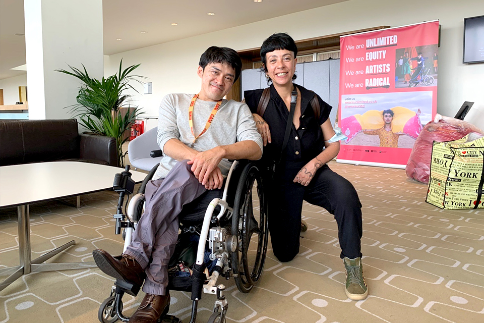 행사장 실내, 휠체어를 탄 김원영 작가와 그 옆에 반무릎으로 앉은 에스텔라 라포니의 기념사진.