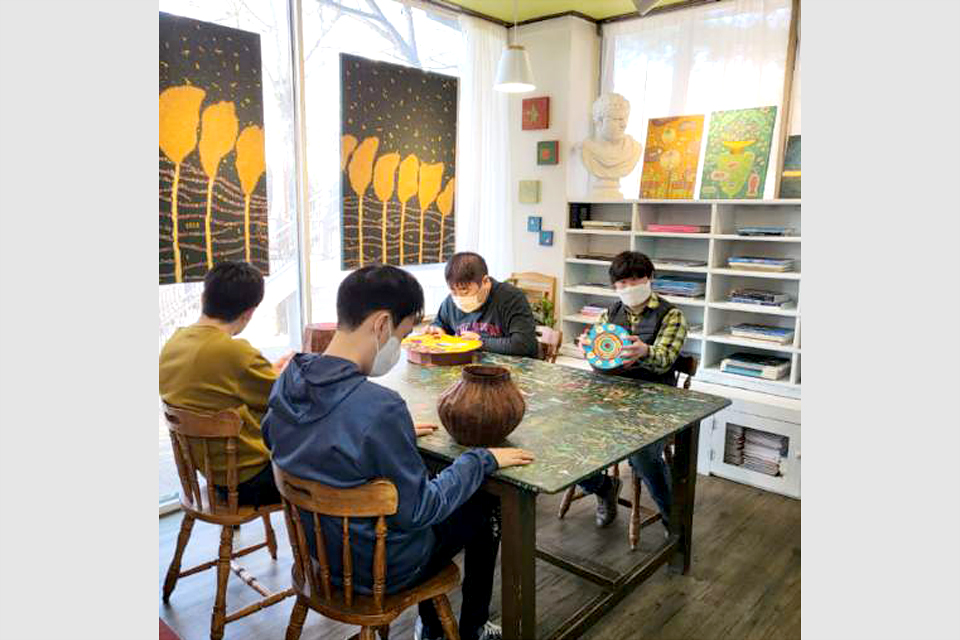 캔버스와 조형물이 잘 정돈된 작업실, 통 유리창 옆 테이블에 앉은 네 명이 각자 조형물을 만들고 있다. 