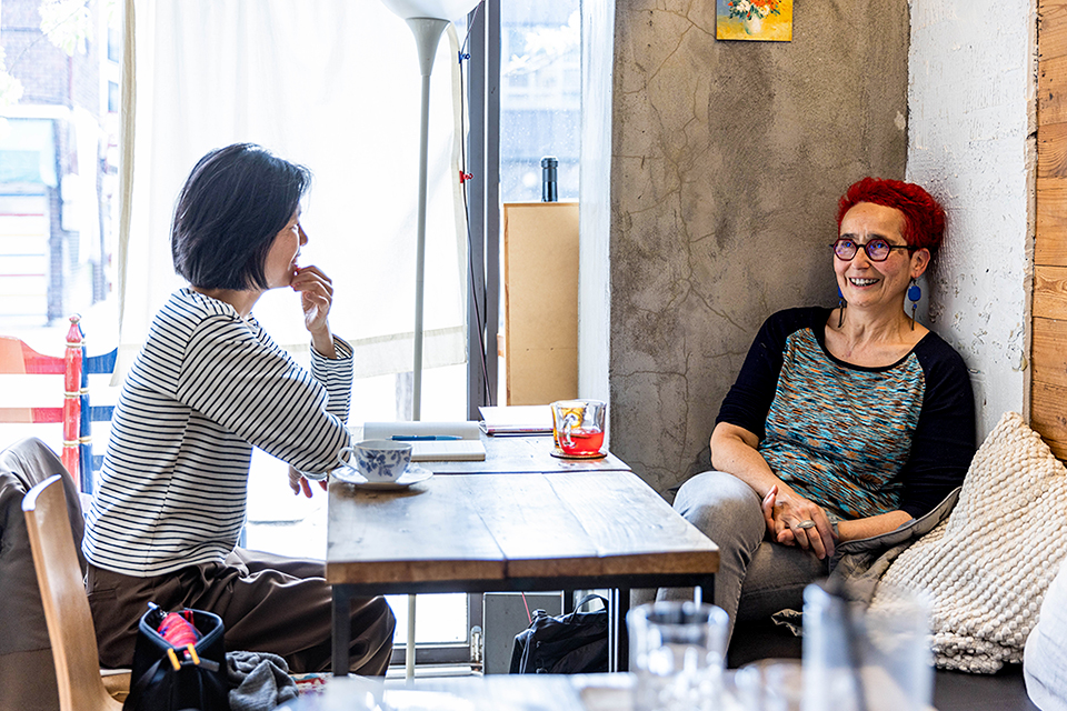 콘크리트 벽체와 나무 테이블로 꾸며진 카페 안. 두 사람이 테이블을 사이에 두고 앉아 웃으며 이야기하고 있다.