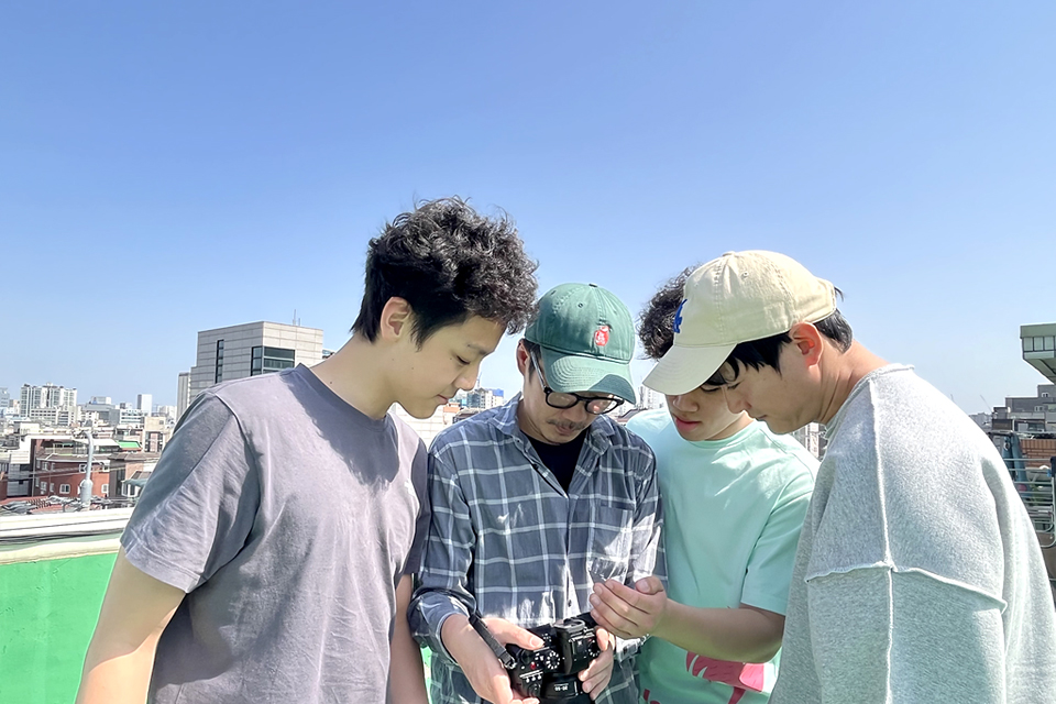 파란 하늘이 보이는 야외. 농청년 셋과 사진작가가 카메라 뷰파인더를 같이 살펴보고 있다.