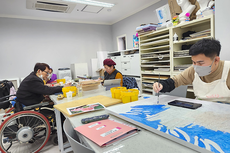 다양한 미술도구들이 잘 정리돼있는 공간, 휠체어장애인 등 네 명이 각자의 그림도구와 캔버스를 테이블 위에 놓고 작업하고 있다.