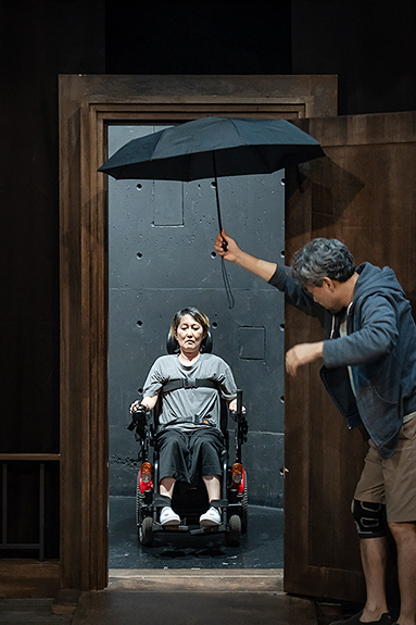 문밖에 휠체어를 탄 여자가 있고 문 안의 남자가 손을 뻗어 우산을 받쳐주고 있다. 