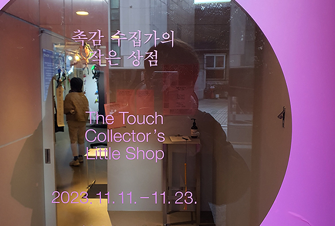 유리문에 보라색 필름을 붙이고, 가운데 동그랗게 비운 부분에는 “촉감 수집가의 작은 상점 The Touch Collector’s Little Shop 2023.11.11.~11.23. 11:00~18:00”이라고 썼다. 유리에 사진 찍는 사람도 비친다.