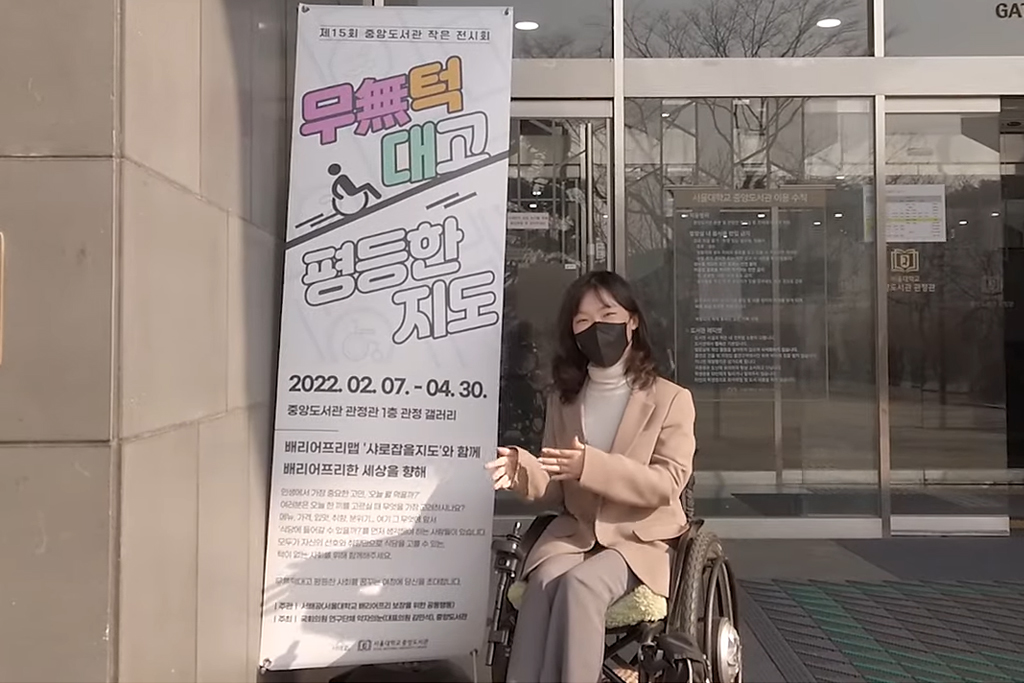 굴러라 구르님 유튜브 영상 캡처. 무턱대고 평등한 지도라고 써있는 행사 배너 앞에 휠체어를 탄 굴러라 구르님이 있다.