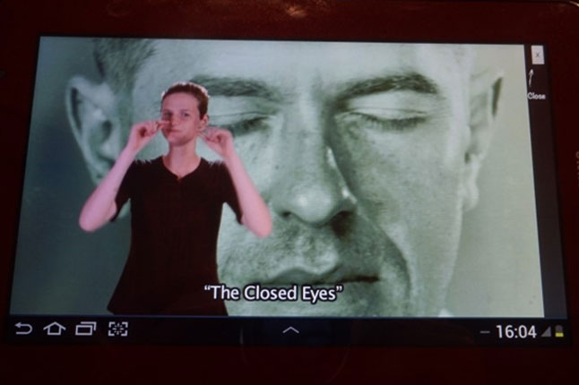 벨기에왕립미술관의 수어해설 비디오 가이드 패드. 작품을 배경으로 왼편에 수어해설사 모습. '눈을 감았다'를 설명하고 있다.
