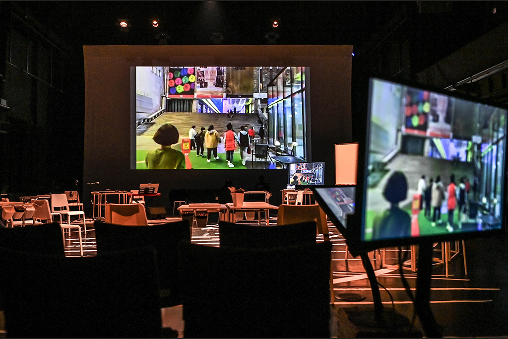 어두운 공연장 안에 수십개의 책상과 의자가 원을 그리며 놓여있다. 뒤쪽 큰 스크린과 무대 가에 있는 프롬프터에는, 공연장 밖을 나가는 많은 사람들의 뒷모습이 담긴 영상이 동시에 보인다. 
