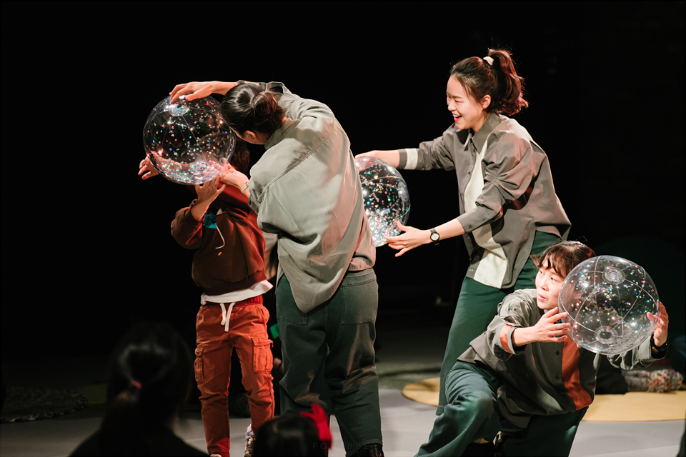 배우 세 명과 관객 어린이 한 명이 반짝이는 커다란 투명 공을 각자 귀에 가져다대고 있다. 