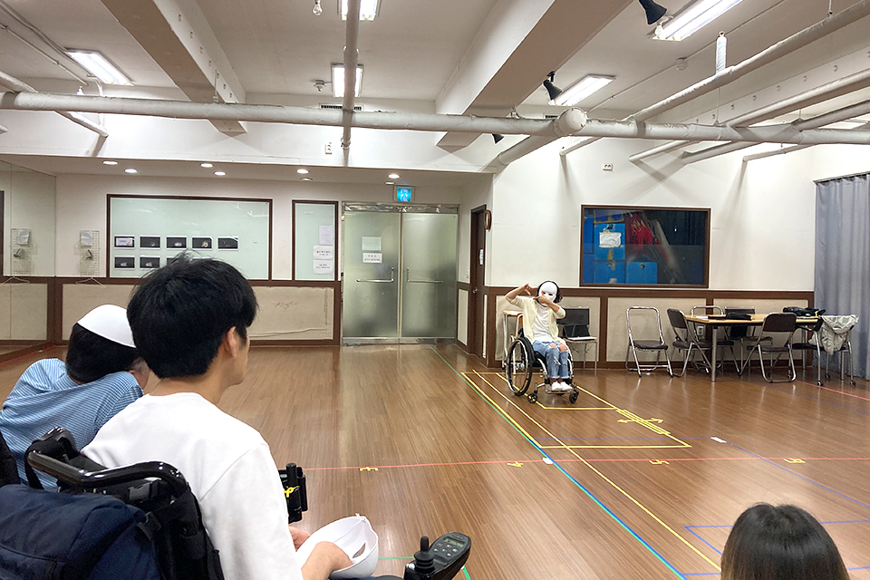 연습실 마룻바닥에 여러 색상 마스킹 테이프로 구획되어 있다. 노란 사각형 안에서 가면을 쓰고 휠체어를 탄 배우가 동작을 취하는 모습을 다른 배우들이 지켜보고 있다.