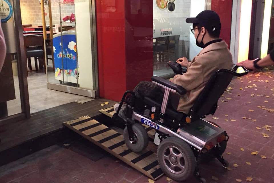 휠체어를 탄 필자가 식당 출입문에 놓인 이동식 경사로로 이동하고 있다.