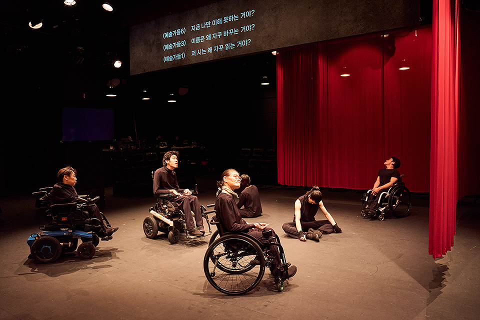 휠체어를 탄 배우들과 바닥에 주저앉은 배우들이 각자 다른 방향을 향해 있다. 한쪽에는 무대와 객석 사이에 붉은 커튼이 드리워져 있다.