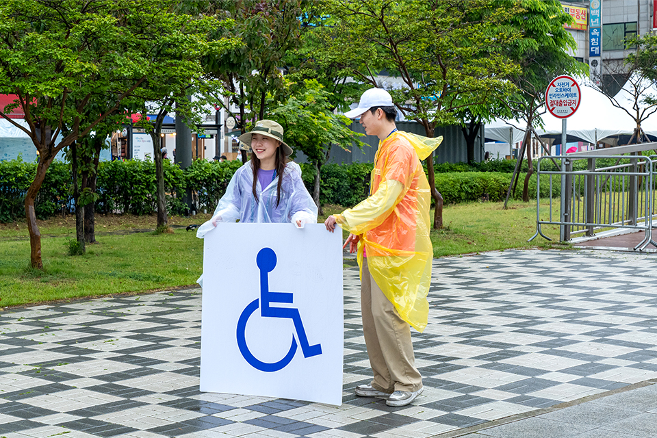 광장에서 우비를 입은 두 사람이 휠체어가 그려진 커다란 패널을 붙들고 서 있다.