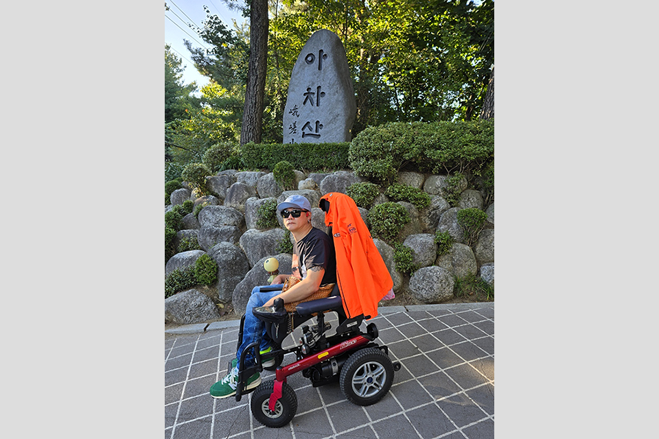 전동휠체어를 탄 필자가 ‘아차산’이라고 쓰여있는 표지석 앞에 있다.