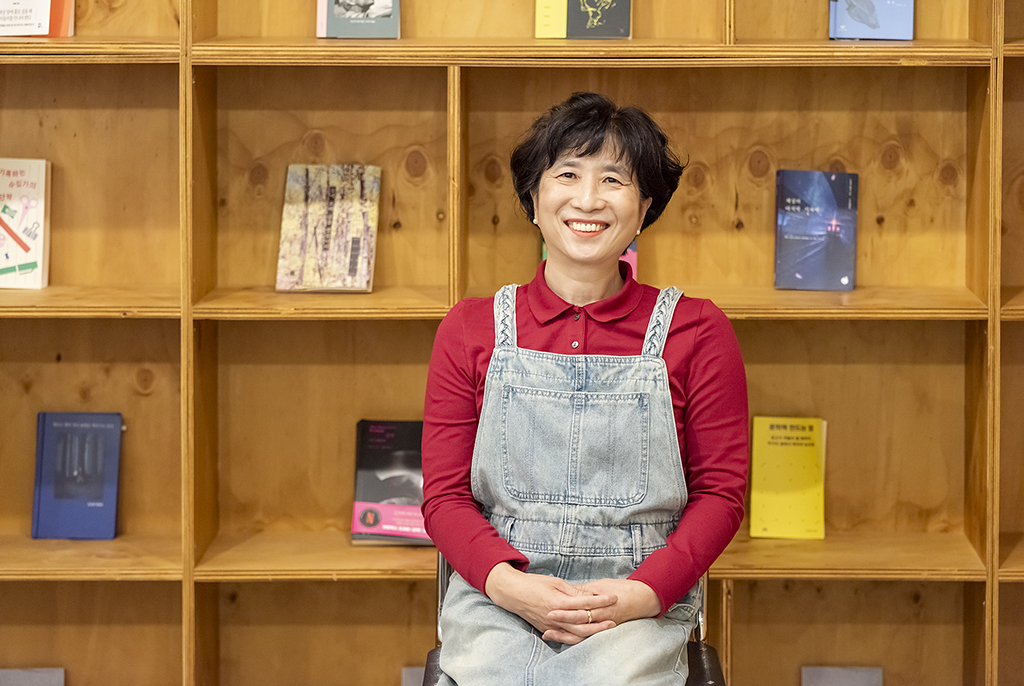 이선영 작가가 책이 진열되어 있는 나무로 짠 전면책장을 배경으로 앉아 환하게 미소짓고 있다.