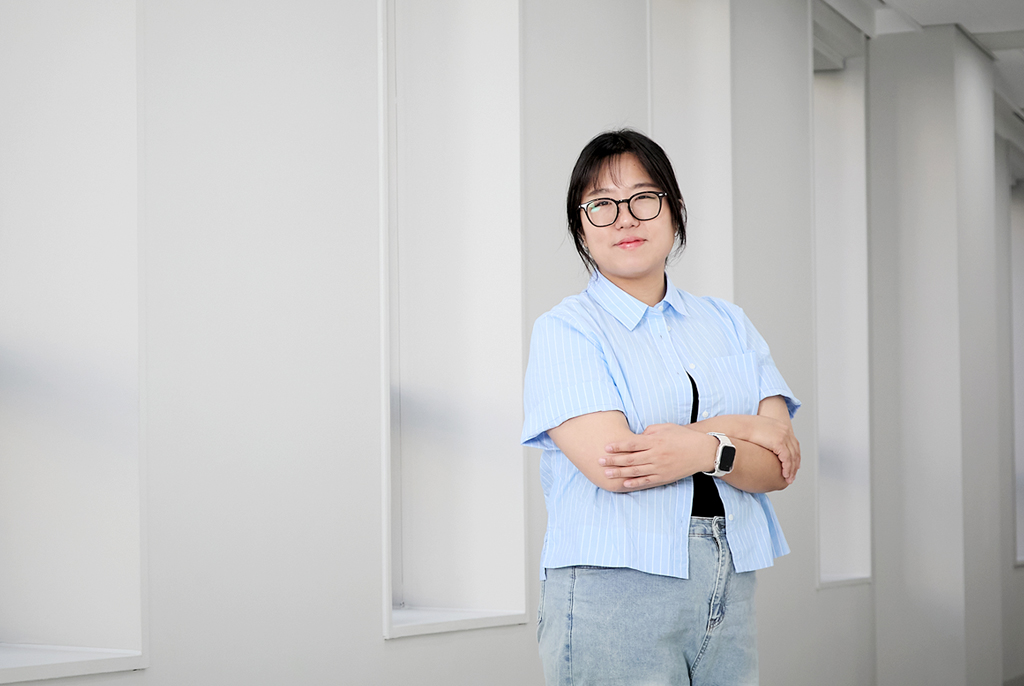 세로로 긴 창문이 줄지어 나 있는 하얀색 복도에서 김은설 작가가 팔짱을 끼고 미소를 띈 얼굴로 카메라를 보고 있다.