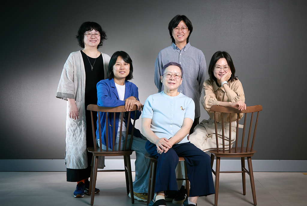 다섯 명이 진한 회색 배경 앞에서 정면을 바라보며 미소짓고 있다. 세 명은 등받이가 있는 의자에 앉아있고, 두 명은 뒷줄에 서 있다.