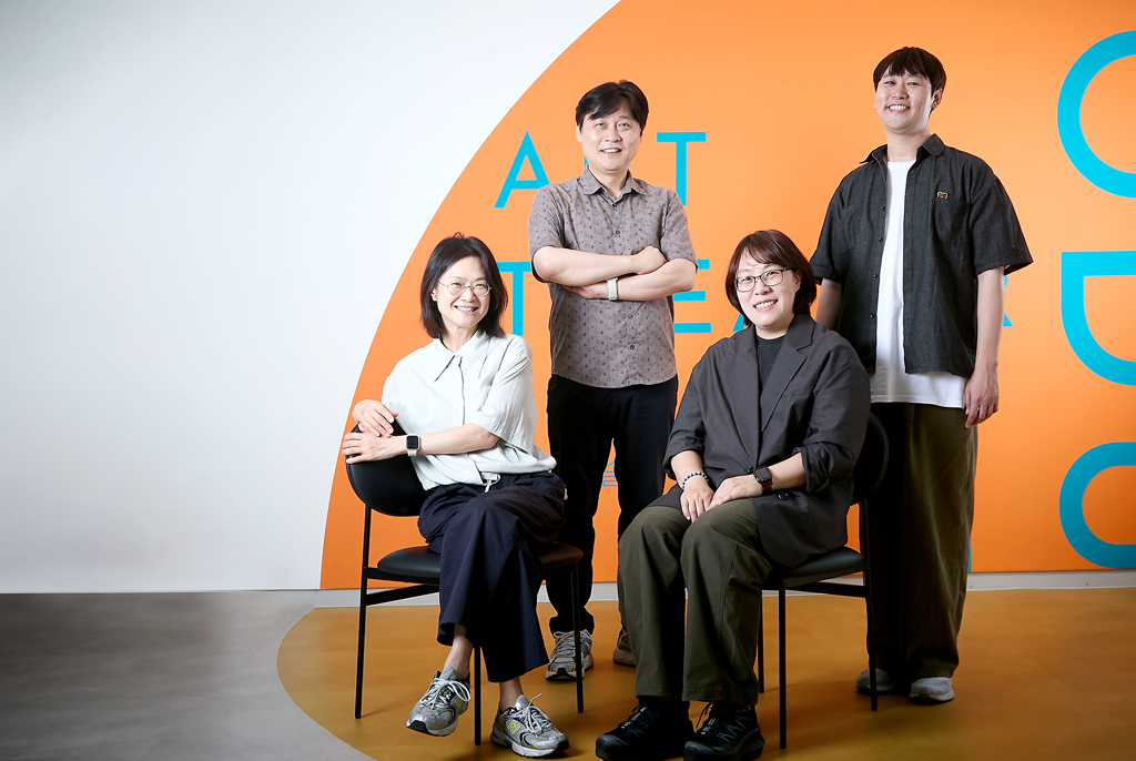 앞줄에 홍은지, 이희원 기획위원이 등받이 의자에 앉아 있고, 뒷줄에 오세형 부장, 이성수 기획위원이 서 있다. 뒷면 흰색 벽에는 주황색으로 칠한 반원에 파란색으로 “ART THEATRE
MODU”라고 쓰여 있는 글자가 일부 보인다.