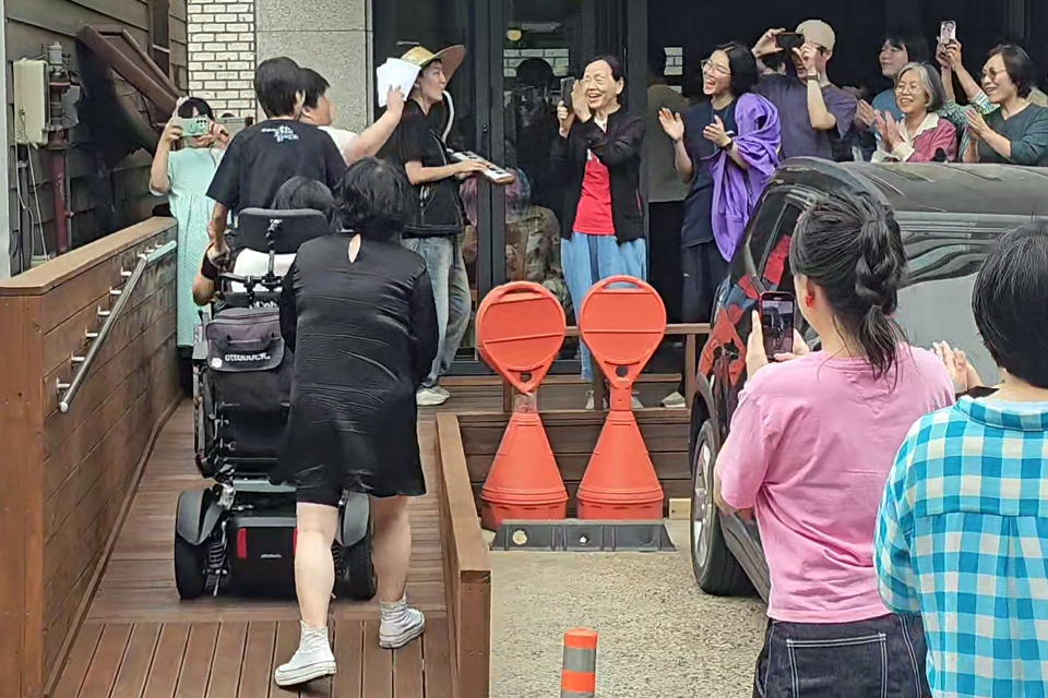 완성된 나무 경사로 위로 전동 휠체어 이용자가 진입하고, 주변 사람들은 박수와 환호로 맞이하고 있다.