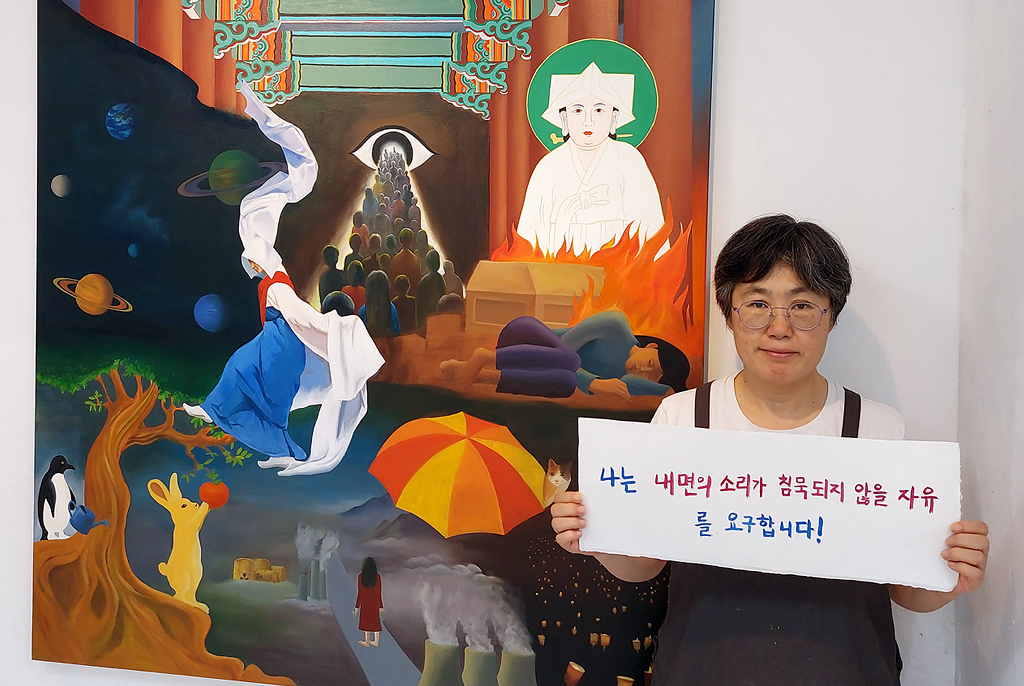 필자가 벽에 걸린 작품 앞에서 ‘나는 내면의 소리가 침묵 당하지 않을 자유를 요구합니다!’라고 적힌 팻말을 들고 서 있다.