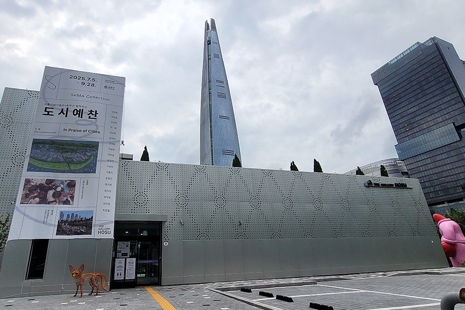 서울시립미술관 SeMA 컬렉션 전시 《도시예찬》이 열리는 더 갤러리 호수 외관. 전시장 입구 옆에는 전시 안내 배너가 세워져 있고, 뒤편으로 롯데월드타워와 고층 건물이 보인다.