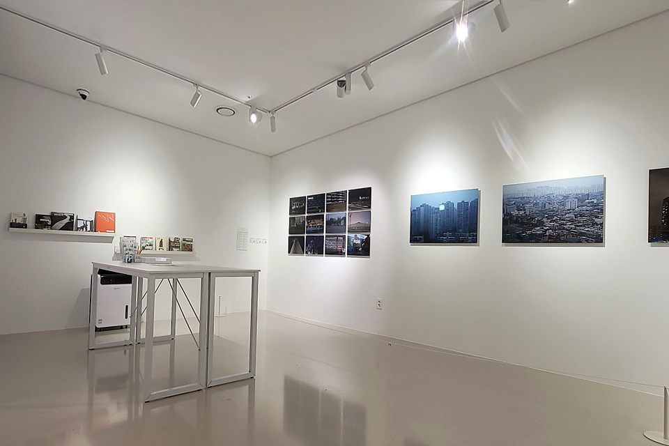 하얀 전시장 벽에 도시 풍경을 담은 사진들이 걸려 있고, 한쪽에는 책과 전시 자료가 진열되어 있다. 가운데에는 흰 테이블이 놓여 있다.