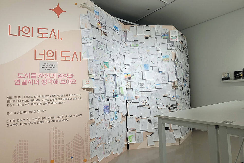 전시장 한쪽 벽면에 ‘나의 도시, 너의 도시’라는 문구와 함께 관람객이 써 붙인 다양한 메모와 그림이 가득 붙어 있다. 옆 테이블 위에는 전시 안내물이 놓여 있다.