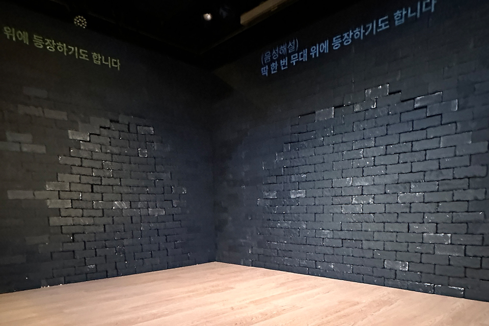 검은 벽과 나무 마룻바닥으로 이루어진 빈 무대 공간. 벽 위에는 자막으로 “(음성해설) 딱 한 번 무대 위에 등장하기도 합니다”라고 투사되어 있다.