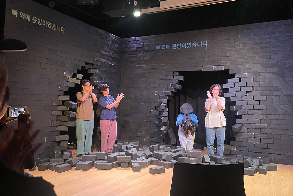 검은 벽돌로 된 무대 세트의 크게 뚫린 구멍 사이로 배우들이 나와 무대 인사를 하고 있다. 바닥에는 벽돌 모양 소품이 흩어져 있고, 세 명은 웃으며 박수치고, 한 명은 고개 숙여 인사하고 있다. 무대 위 벽에는 “빠 역에 윤방이었습니다”라는 자막이 투사되어 있다. 객석에서 관객들이 박수치며 이 장면을 지켜본다.