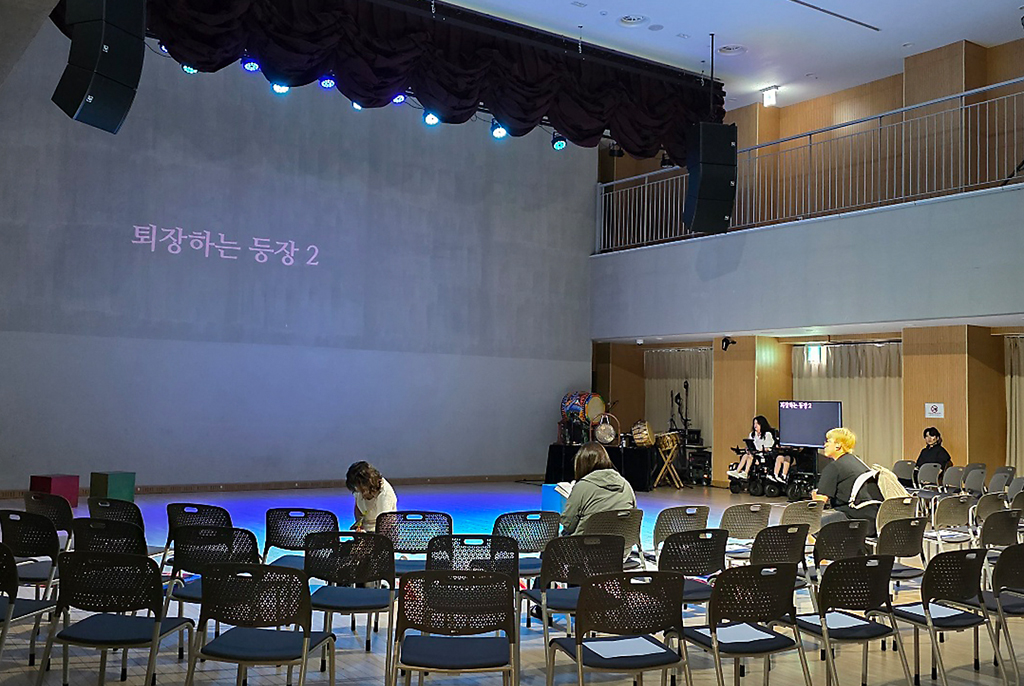 공연을 준비하는 모습. 무대 위 스크린에 ‘퇴장하는 등장 2’라는 글자가 비친다. 객석에는 접이식 의자가 깔려 있고, 몇 명의 관객이 흩어져 앉아 있다.