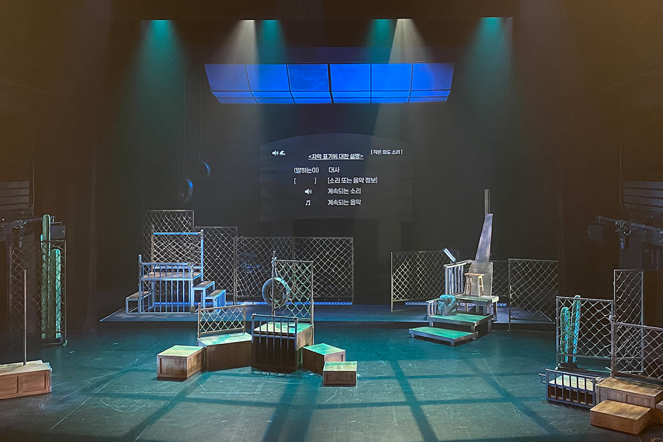 철망과 나무 구조물이 설치된 무대. 중앙 스크린에는 대사, 소리, 음악에 대한 자막 설명이 표시되어 있다.