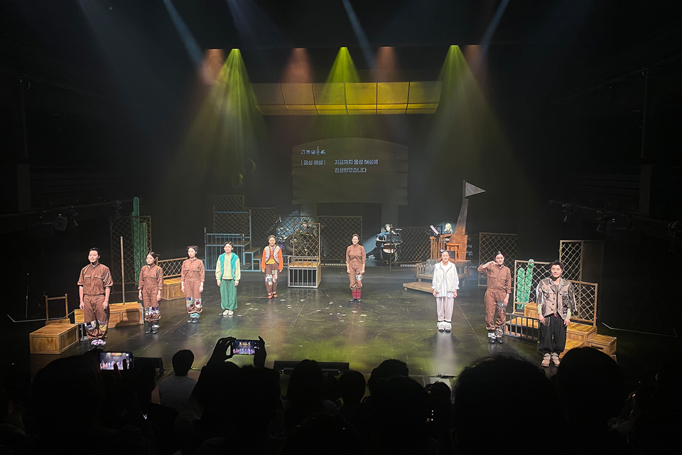 무대 위에 여러 명의 배우가 나란히 서서 관객을 향해 인사하고 있다. 뒤편에는 철망과 나무 구조물, 악기가 놓여 있다.