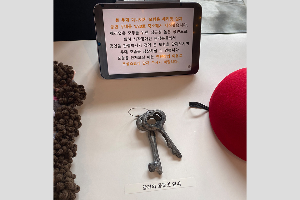 테이블 위에 태블릿 안내문과 금속 열쇠 모형이 놓여 있다. 안내문은 무대 미니어처 모형이 실제 공연 무대를 축소 제작한 것임을 설명하며, 아래에는 ‘찰리의 동물원 열쇠’라는 설명을 붙였다.