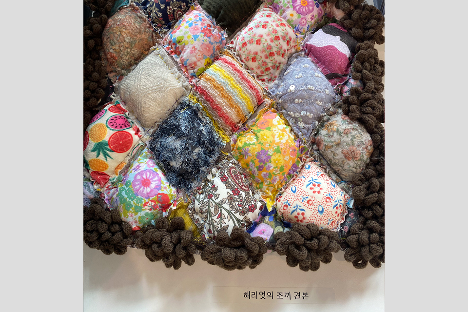 다양한 색과 무늬의 천 조각을 이어 만든 작은 패치워크 작품. 네모난 조각마다 솜이 들어 있어 입체적으로 볼록하게 솟아 있으며, 가장자리는 갈색 털실로 장식되어 있다. 아래에는 ‘해리엇의 조끼 견본’이라는 설명을 붙였다.