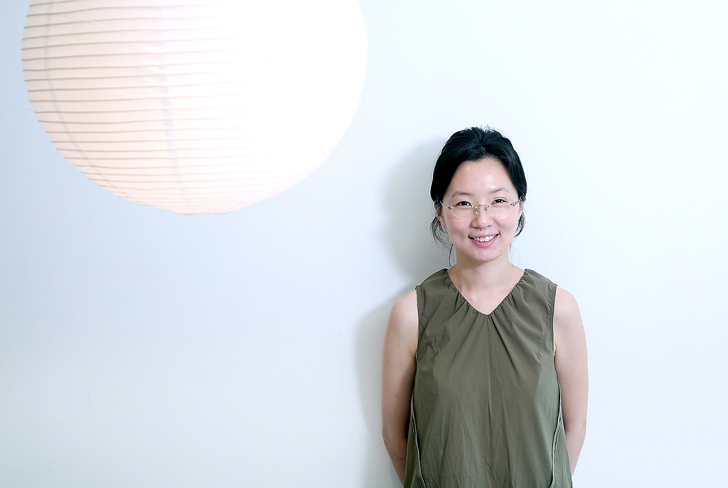 흰 벽 앞에 선 김보라 작가가 두 손을 뒤로 모으고 서서 정면을 바라보며 미소 짓고 있다. 왼쪽 위에는 둥근 종이 조명등이 걸려 있다.