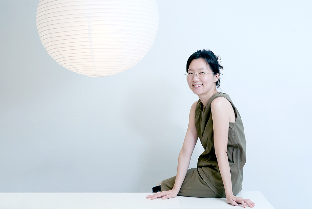 흰 벽 앞에서 김보라 작가가 흰색 벤치의자에 비스듬하게 걸터앉은 채 정면으로 고개를 돌려 미소 짓고 있다. 왼쪽 위에는 둥근 종이 조명등이 걸려 있다.