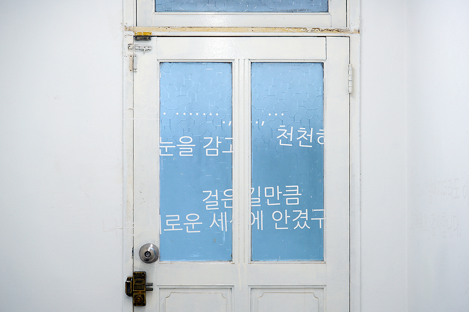 하얀색 벽과 나무문 유리창에 흐릿하게 흰색 문구가 새겨져 있고, 파란빛이 비치는 유리 위에 “눈을 감고… 천천히… 걸은 길만큼… 새로운 세계에 안겼구…”라고 쓰여 있는 일부 글자가 읽힌다.