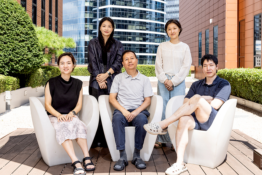 야외 정원에 놓인 흰색 의자에 앉거나 서서 포즈를 취한 다섯 명이 카메라를 향해 있다. 뒤편으로는 잘 다듬어진 녹색 조경과 현대적인 고층 빌딩 외관이 보인다.