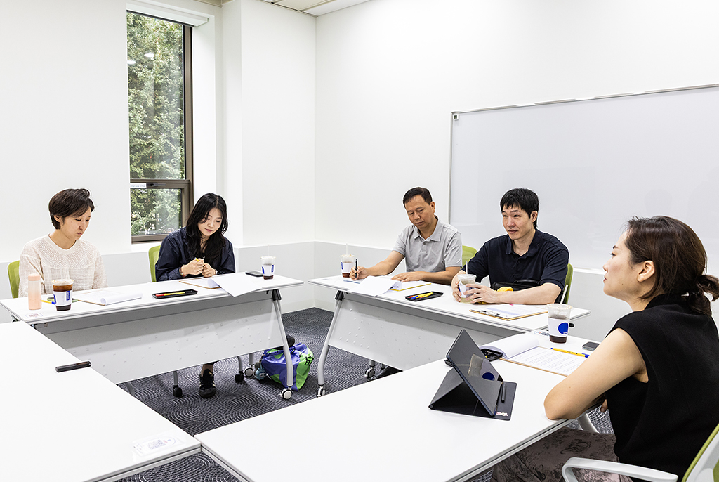 흰 벽으로 둘러싸인 회의실에서 다섯 명이 ㅁ자 형태로 배치된 책상에 둘러앉아 회의 중이다. 책상 위에는 회의자료와 음료가 놓여 있고, 벽에는 큰 화이트보드와 창문이 보인다.