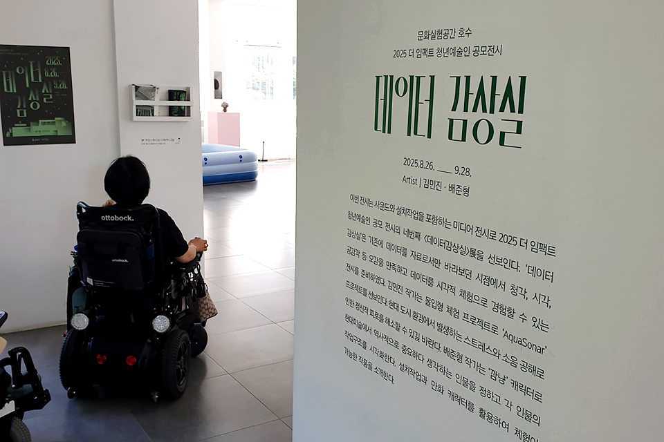 전시장 입구의 하얀 벽에 전시 안내문이 적혀 있고, 그 옆으로 휠체어를 탄 관람객이 전시장 안으로 들어가고 있다. 전시 안내문에는 전시 제목 《데이터 감상실》과 전시 일정, 참여 작가 김민지·배준형의 이름이 적혀 있다. 