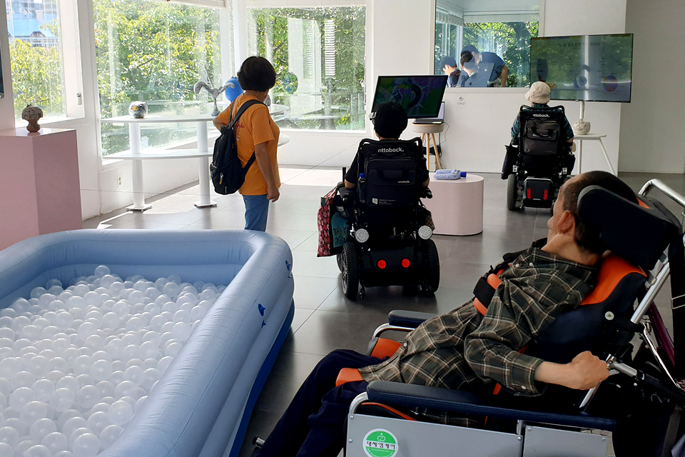 전시장 내부 전경. 넓은 유리창을 통해 자연광이 들어오는 밝은 공간이다. 앞쪽에는 흰색 공이 가득 찬 푸른색 에어풀이 설치되어 있고, 그 뒤로 두 대의 모니터가 전시작품 영상을 재생 중이다. 보행인 한 명과 전동휠체어를 탄 세 명의 관람객이 전시를 감상하고 있다. 