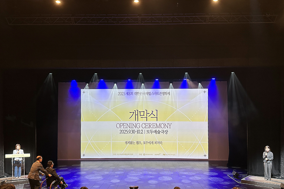 무대 중앙 대형 스크린에 ‘2025 제8회 대한민국 패럴스마트폰영화제 개막식(Opening Ceremony) 2025.9.30–10.2 모두예술극장’이라는 문구가 크게 영사되고 있다. 왼쪽에는 연단 앞에 사회자가 서 있고, 오른쪽에는 검은 옷을 입은 수어통역사가 서 있다. 무대 양측 모니터에는 문자통역 자막이 표시되고 있다. 객석 앞쪽에는 휠체어 이용자들이 앉아 있다. 