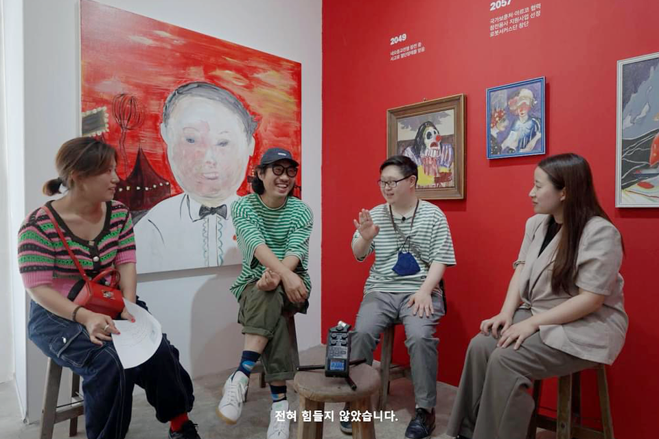 붉은색 벽면에 인물화와 서커스를 연상시키는 그림들이 걸린 전시 공간에서 네 사람이 나란히 앉아 얼굴을 보며 대화 나누고 있다. 화면 하단에는 자막으로 “전혀 힘들지 않았습니다.”라고 쓰여 있다.