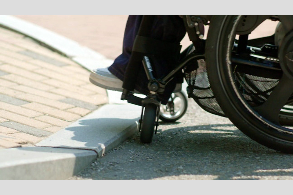 휠체어 앞바퀴가 인도 턱을 오르려는 순간이 클로즈업되어 있다. 회색 도로와 연석, 벽돌길의 경계가 도드라져 보인다.