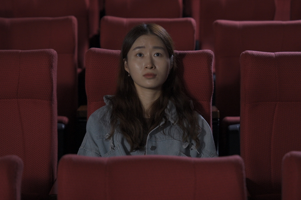 붉은 좌석이 줄지어 있는 어두운 영화관에서 한 여성이 중앙에 혼자 앉아 정면을 바라보고 있다.