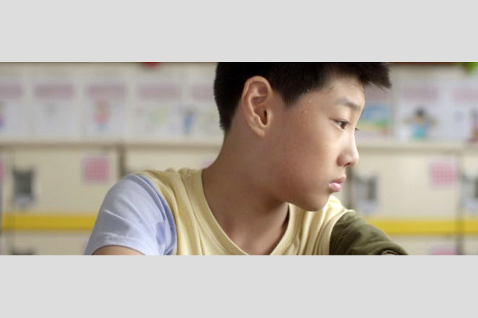 교실에 한 소년이 고개를 옆으로 돌린 채 생각에 잠긴 듯 앉아 있다. 뒤쪽 벽에는 학생들의 그림과 글이 붙어 있다.