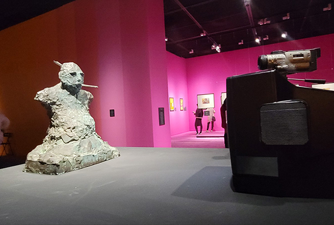 강렬한 분홍색 조명과 벽면이 구성된 전시 공간에 거칠게 조각된 흉상 형태의 조형물이 전경에 놓여있고, 오른쪽에는 오래된 캠코더가 설치된 장치가 보인다. 뒤편 전시실에는 관람객들이 벽에 걸린 회화 작품을 보고 있다.