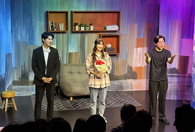 커튼콜 중인 세 배우가 객석을 향해 서서 관객에게 인사하고 있다. 왼쪽은 검은 정장 차림, 가운데는 체크 셔츠와 청바지를 입고 꽃다발을 들고 있으며, 오른쪽은 검은 티셔츠와 바지를 입고 있다. 