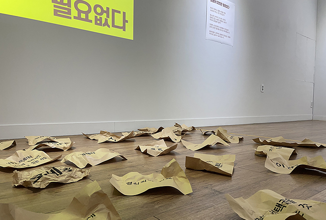 전시장 바닥에 구겨진 갈색 종이들이 흩어져 놓여있다. 각각의 종이에는 검은 글씨로 다양한 문장이 적혀 있다. 벽면에는 노란 바탕에 ‘필요없다’라는 문구가 크게 투사돼 있으며, 오른쪽에는 작품 설명 텍스트가 게시되어 있다.